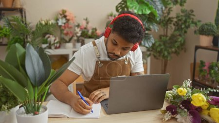 A focused african man in apron using laptop and headphones while taking notes surrounded by plants in a flower shop.