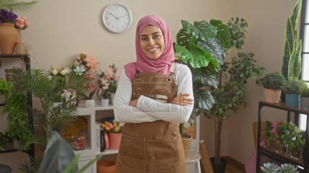 Téléchargez les photos : Une femme souriante aux bras croisés portant un hijab et un tablier se tient dans un salon rempli de plantes. - en image libre de droit