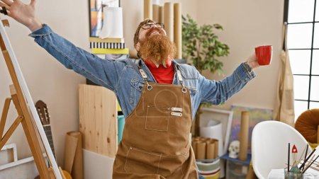 Photo for Confident young redhead man artist, gleefully drinking coffee while flashing a hearty smile amidst his art studio. - Royalty Free Image