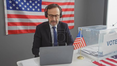 A mature man wearing headphones sits at a laptop in a voting center with an american flag and ballot box.