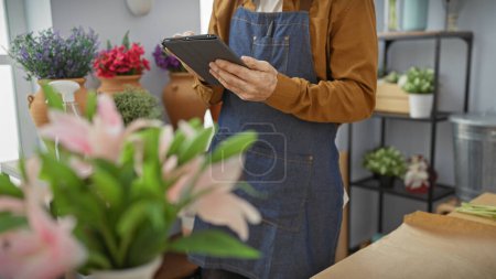 Téléchargez les photos : L'homme hispanique à la barbe utilise une tablette dans un magasin de fleurs entouré de plantes assorties, donnant une ambiance d'entrepreneur moderne. - en image libre de droit