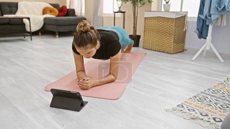 Mujer hispana joven y deportiva que se dedica a un entrenamiento serio en línea en casa, convirtiendo su sala de estar en un centro de fitness con su computadora portátil. 