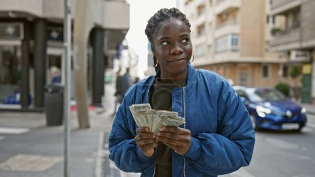 Foto de Mujer africana con chaqueta azul cuenta con dinero en una calle urbana concurrida, mostrando la planificación financiera en un entorno urbano. - Imagen libre de derechos