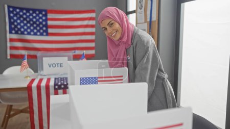 A smiling woman with a hijab votes in a room adorned with american flags, representing democracy and diversity.