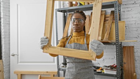 Foto de Mujer afroamericana con trenzas con gafas de seguridad y guantes sosteniendo una montura de madera en un taller. - Imagen libre de derechos