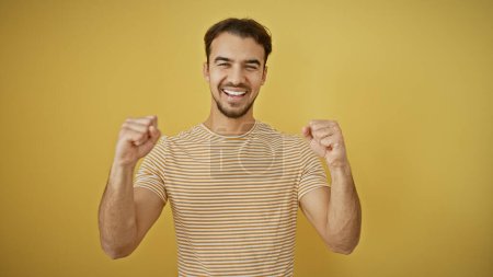 Téléchargez les photos : Jeune homme hispanique à la barbe souriante et levant les poings avec succès sur un fond jaune. - en image libre de droit