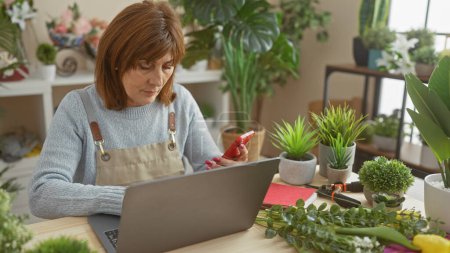 Mature woman using smartphone and laptop at florist shop amidst green plants in indoor setting