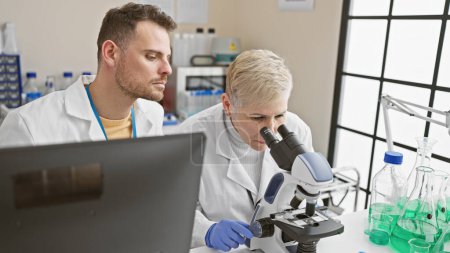 A woman and man in lab coats work together, analyzing samples with a microscope in an indoor laboratory.