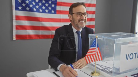 Smiling mature man in suit casting ballot with american flag backdrop inside voting center.