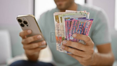 Young man sitting at home on the living room sofa, banking business at his fingertips, holding a stack of crisp hong kong dollar banknotes, glued to his brightly lit smartphone screen.
