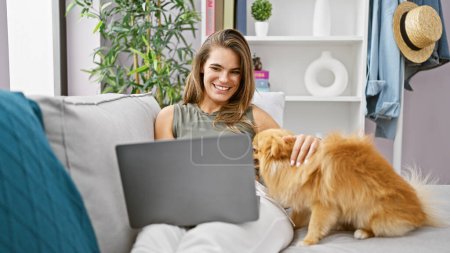 Foto de Mujer hispana alegre y joven sentada en el sofá, usando el portátil con su perro juguetón, disfrutando de la tecnología en línea en la comodidad de su sala de estar en casa - Imagen libre de derechos