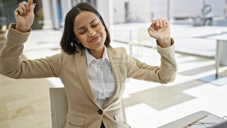 Foto de Una mujer hispana alegre, estirándose en la oficina moderna con una chaqueta beige, irradia satisfacción y relajación en un entorno profesional. - Imagen libre de derechos