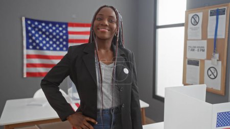 Confident african american woman with braids wearing voter sticker, posing in a polling station with us flag.