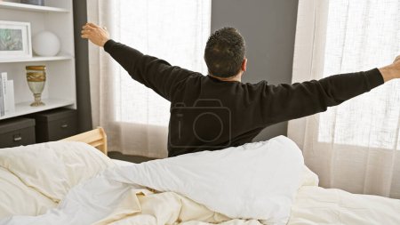 A young man stretches upon waking in a well-lit, cozy bedroom setting, exuding a sense of morning vitality and comfort.