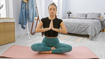 Foto de Mujer hispana joven y hermosa y serena sumergiéndose en una tranquila sesión de yoga, sentada en el suelo de su acogedor dormitorio, cultivando la calma y el equilibrio a través de su estilo de vida fitness. - Imagen libre de derechos