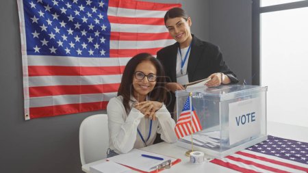 Two smiling women conducting electoral process in an american college with a prominent flag and voting box in an indoor setting.