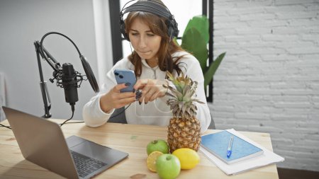 Young woman with headphones in a radio studio looking at phone by microphone and laptop, evoking a creative workspace.
