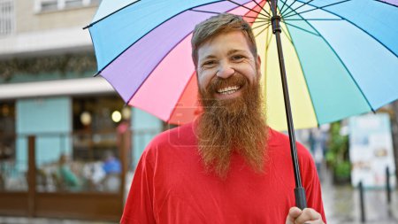 Foto de Joven pelirrojo sonriendo confiado sosteniendo paraguas en la calle - Imagen libre de derechos