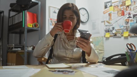 Hispanic woman detective sips coffee and examines phone in cluttered police station office, surrounded by evidence.