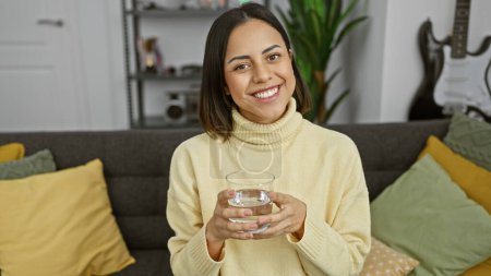Photo for A cheerful woman in a cozy living room drinking water, exuding warmth and wellbeing. - Royalty Free Image