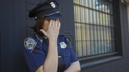 Téléchargez les photos : Une policière affligée en uniforme debout près d'un immeuble de la ville, couvrant son visage de sa main. - en image libre de droit