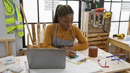 Focused african woman with braids planning in a carpentry workspace, surrounded by tools and safety equipment.