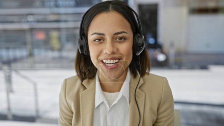 Téléchargez les photos : Femme hispanique souriante avec écouteurs dans un costume beige sur fond de bureau flou. - en image libre de droit