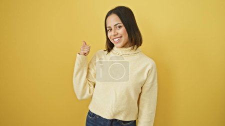 Photo for Smiling young hispanic woman gesturing thumbs up against a vibrant yellow wall, exuding positivity and confidence. - Royalty Free Image