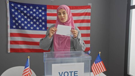 Eine junge hispanische Frau mit Hidschab wählt drinnen vor dem Hintergrund amerikanischer Flaggen.