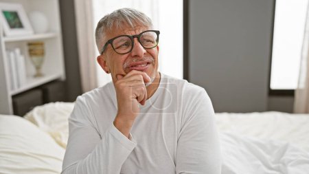Ein nachdenklicher Mann mittleren Alters mit grauen Haaren und Brille sitzt drinnen auf einem Bett, die Hand am Kinn und betrachtet.