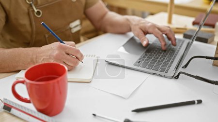 Photo for A middle-aged man sketches in a notepad while using a laptop in his woodworking workshop. - Royalty Free Image
