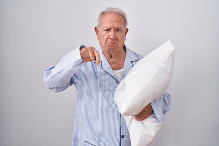 Hombre mayor con el pelo gris usando pijama abrazando almohada apuntando hacia abajo mirando triste y molesto, indicando la dirección con los dedos, infeliz y deprimido. 