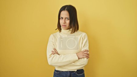 Young hispanic woman with crossed arms looking annoyed against a yellow wall.
