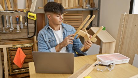 Foto de Hermoso joven carpintero hispano trabajando en un objeto de madera en el taller de carpintería, navegando hábilmente por Internet en su computadora portátil. - Imagen libre de derechos
