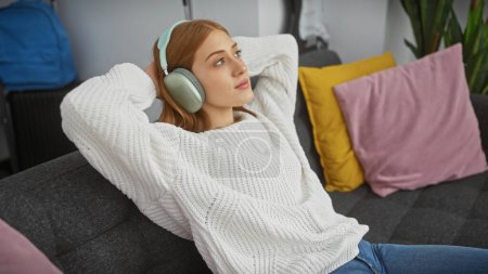 Téléchargez les photos : Femme rousse se détend à la maison sur le canapé, écouter de la musique avec des écouteurs dans un salon confortable. - en image libre de droit