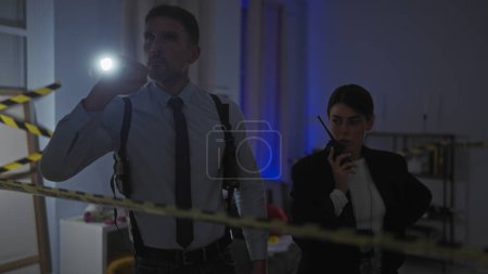 A man and woman investigating a crime scene indoors with caution tape and flashlights at night.