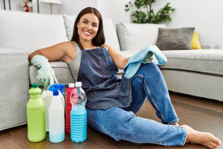 Téléchargez les photos : Jeune belle femme hispanique souriant confiant assis sur le sol avec des produits propres à la maison - en image libre de droit
