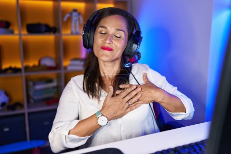 Brünette Frau mittleren Alters, die Videospiele spielt, lächelt mit den Händen auf der Brust, die Augen geschlossen mit dankbarer Geste im Gesicht. Gesundheitskonzept. 