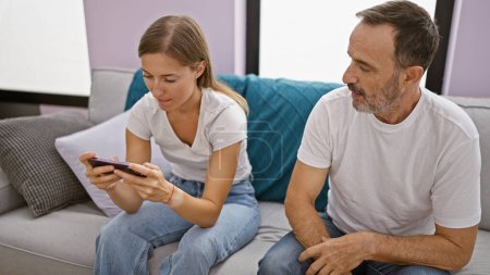 Téléchargez les photos : Père et fille assis sur le canapé en utilisant un smartphone à la maison - en image libre de droit