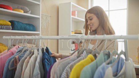 Photo for A smiling young woman casually browses through colorful clothing in a well-lit, neat dressing room. - Royalty Free Image