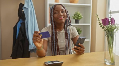 Téléchargez les photos : Femme afro-américaine avec des tresses souriantes, tenant carte de crédit et smartphone à la maison. - en image libre de droit