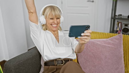 Téléchargez les photos : Femme mûre gaie aux cheveux blonds jouissant de la musique sur écouteurs et prenant un selfie à la maison. - en image libre de droit