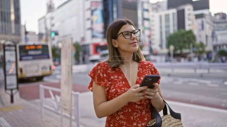Smiling beautiful hispanic woman in glasses uses phone on bustling tokyo street, enjoying modern city's vibe, cheerfully texting away