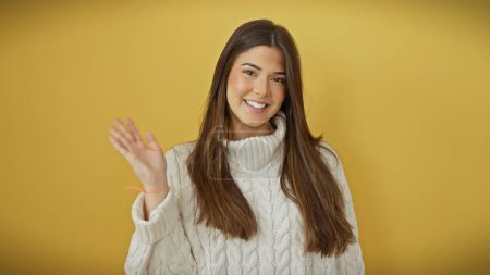 Foto de Mujer joven hispana sonriente usando un suéter blanco aislado sobre un fondo de corte amarillo, irradiando belleza y calidez. - Imagen libre de derechos
