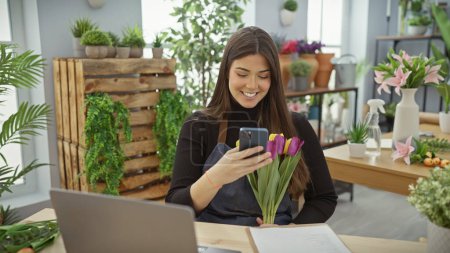 Foto de Mujer sonriente con flores usando teléfono inteligente en una vibrante tienda de flores de interior. - Imagen libre de derechos