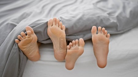 Madre e hija tumbadas juntas, moviendo cómodamente sus pies en una acogedora cama en el dormitorio, compartiendo una mañana relajada en su casa, profundizando el vínculo de su relación en el interior