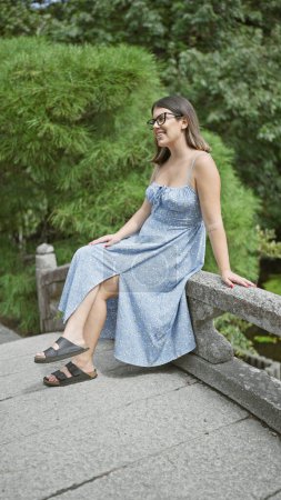 Téléchargez les photos : Belle jeune femme hispanique portant des lunettes souriant assis sur un pont au parc japonais - en image libre de droit