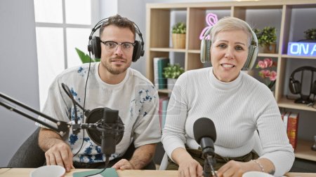 Man and woman wearing headphones speaking into microphones in a bright radio studio with 'on air' sign