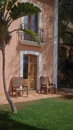 Téléchargez les photos : Charmante porte de villa en terre cuite flanquée de chaises en bois et d'une végétation luxuriante, invitant à la détente et au luxe. - en image libre de droit