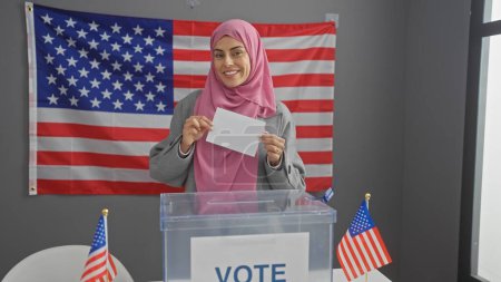 A smiling young hispanic woman wearing a hijab holds a ballot in a college electoral center with american flags.